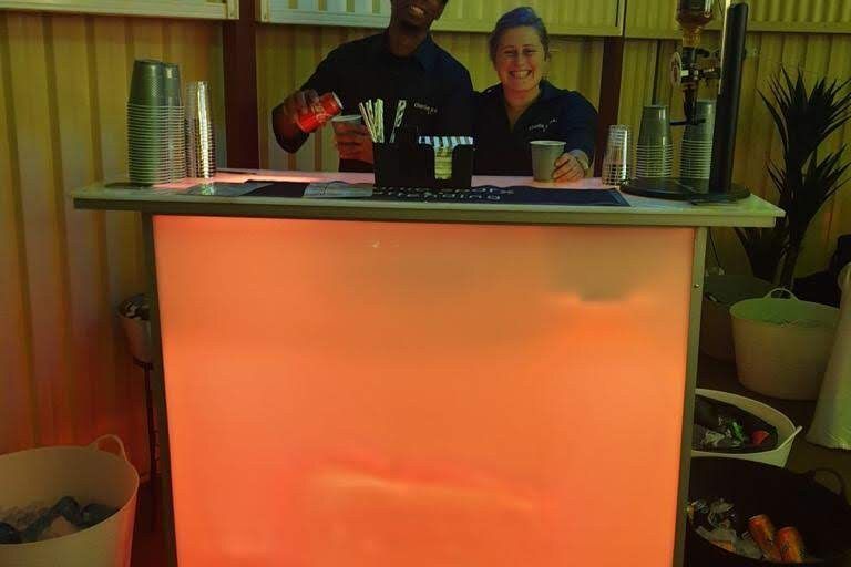 Bartender behind the glow bar lit warm orange at an outdoor evening party