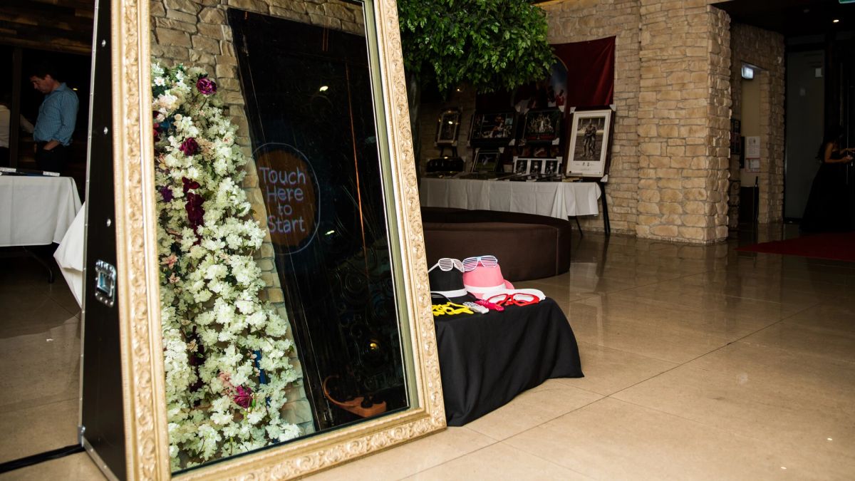 Gold ornate mirror booth with a white and burgundy flower wall and a props table, set up in a stone-walled Sydney venue foyer before guests arrive
