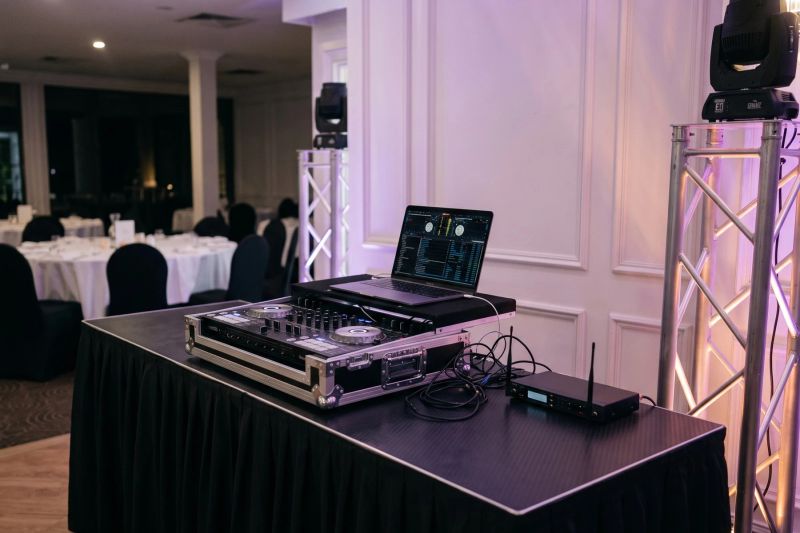 Illustration of a DJ desk with a controller, laptop and wireless microphone receiver beside moving-head lights on a truss stand in a function room