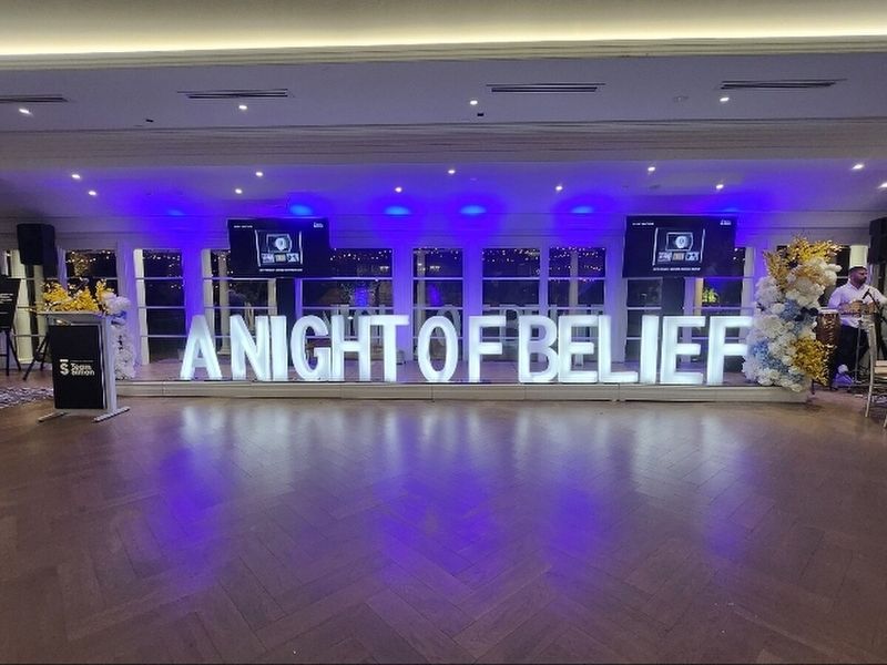 Marquee letters spelling A NIGHT OF BELIEF across a lit dance floor