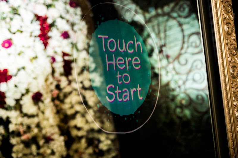 Close-up of the mirror booth touch screen prompting guests to start, framed by the flower wall
